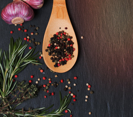 Dark culinary background with a wooden spoon with different pepper, garlic, rosemary and herdsの写真素材