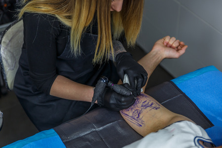 Blonde artist girl carefully draws a tattoo with her tattoo machine, on a boy's armの写真素材