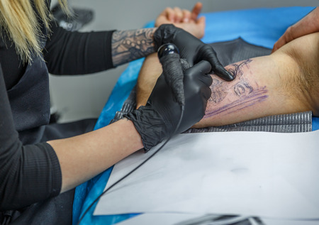 Blonde girl draws a tattoo with her tattoo machine on a boy's armの写真素材