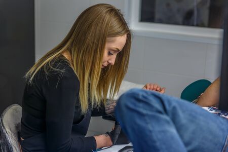 Blonde woman sketches a special tattoo with her tattoo machine on the  arm of a boy
