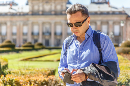 A man in a blue shirt and black glasses checks things on his mobile phone while he is in the middle of a parkの写真素材