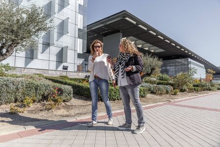 Two businesswomen wearing casual clothes, having a coffee while chatting in front of a large office buildingの写真素材