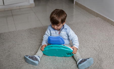 A cute little blond boy examines a toy, while sitting on the carpet in his roomの写真素材