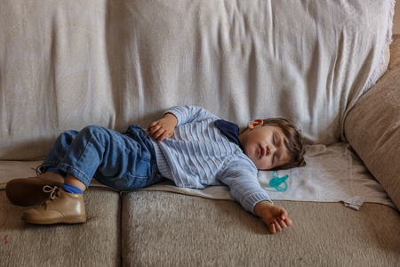 Cute and small boy dressed in blue and brown shoes, is sleeping peacefully on large living room sofa of his homeの写真素材