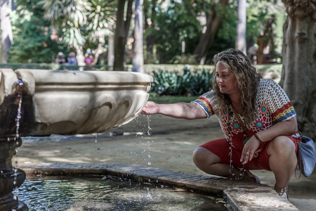 A woman quietly picks up water with her right hand, which springs from a spring in a fountain in a parkの写真素材