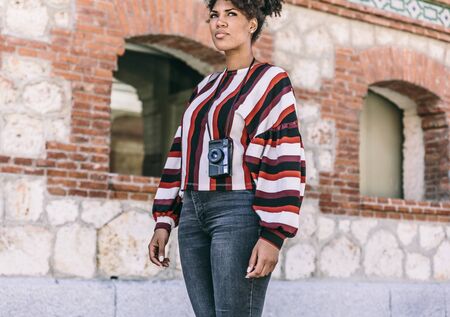 Beautiful dark skinned girl with casual clothes and a vintage camera is standing on a street thinkingの写真素材