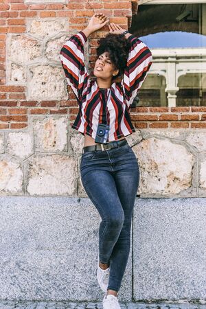 Beautiful tourist girl with dark skin and afro hair leaning on a brick wall next to a window, while visiting a cityの写真素材