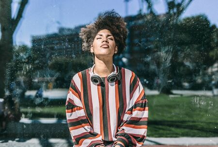 Beautiful woman with afro hair wearing headphones and with closed eyes, is sitting in a large window of an outdoor buildingの写真素材