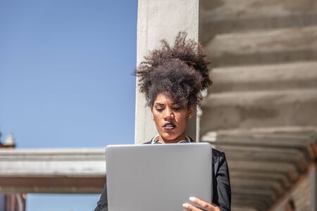 Attractive girl with black jacket, headphones around neck and afro hair, is concentrated with her laptop while leaning on an outdoor column on a sunny dayの写真素材