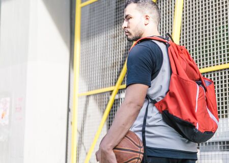 Side view of a sportsman walking through an urban area carrying his basketball and a large red backpack on a sunny afternoonの写真素材