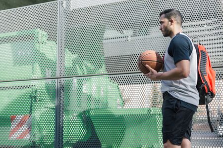 Attractive basketball player walks beside a metal wall carrying his backpack and his orange ball on...