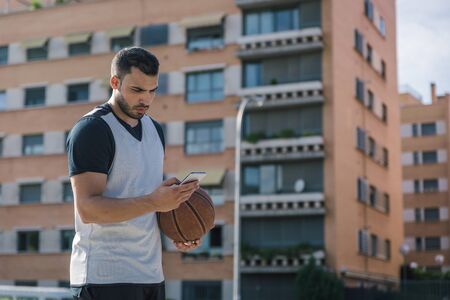 Attractive sportsman man with a basketball looks at his smartphone while taking a breakの写真素材