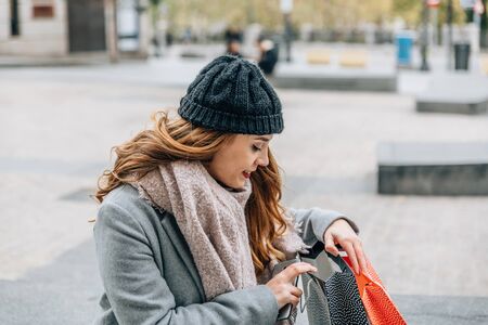 European blonde woman wearing coat, scarf and hat checks her shopping bags while sitting on a bench in the streetの写真素材