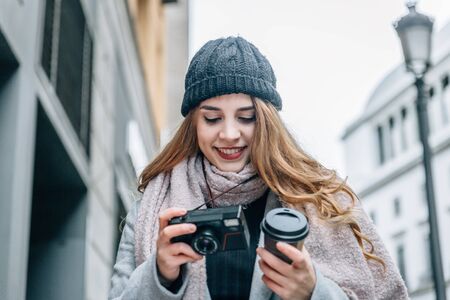 Warm blonde caucasian girl looks at her vintage camera with a cup of coffee in the other hand, walking down the streetの写真素材