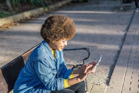Smiling afro haired girl writes on her mobile phone sitting on a bench in a park one autumn dayの写真素材