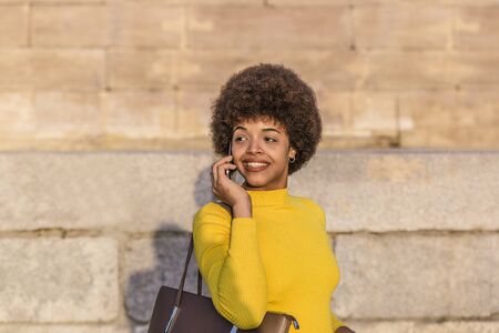 Beautiful mixed race girl with afro hair wearing yellow shirt and a handbag talking on her mobile...