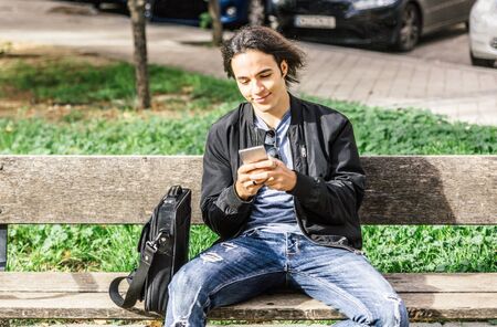 Smiling and happy Caucasian handsome guy looks relaxed at his smartphone sitting on a park bench on...
