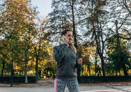 Young blonde woman in sports tracksuit smiles while talking on her smartphone in a park on an autumn dayの写真素材