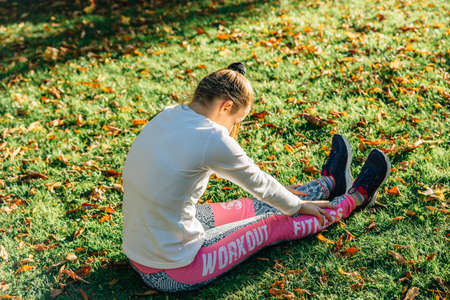 Blonde girl stretches her back sitting on the grass before training on a fall dayの写真素材