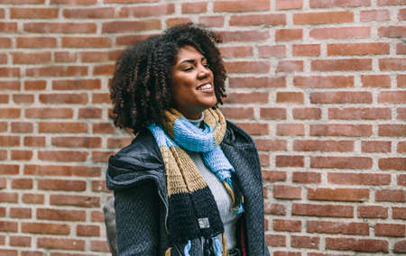 Happy brunette woman in coat and scarf walks smiling in front of a brick building
