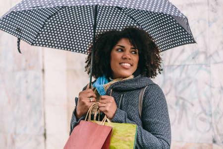 Beautiful afro styled brunette woman smiles with an umbrella and some bags in front of a gray wallの写真素材