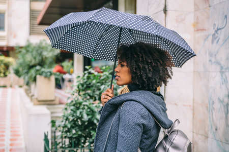 Attractive brunette with afro hair with an umbrella and a coat in front of a buildingの写真素材