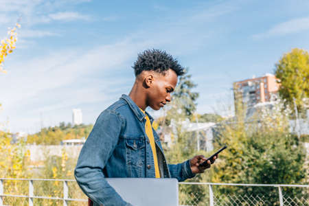 Young boy with afro hair looks worriedly at his mobile phone in a city in a sunny dayの写真素材