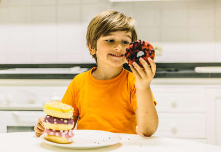 Smiling blond boy looks at a donut in the kitchen at home with orange t-shirtの写真素材