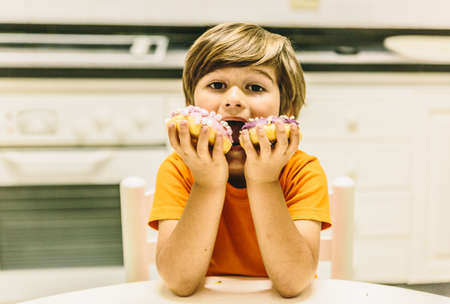 Caucasian blond boy eats two donuts at the same time in the kitchen of his homeの写真素材
