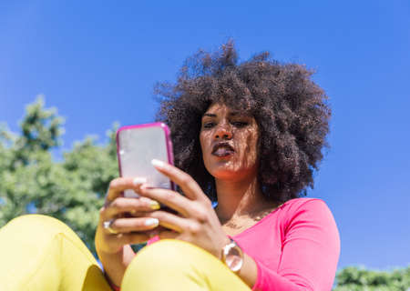 Beautiful racial woman with afro hair looks at her mobile phone outdoors on a sunny day
