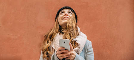 Blonde girl with a smartphone looks up with positivity and happiness in front of a wall in the...