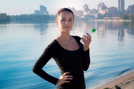 Young pretty fitness woman holds water bottle at morning river and city on backroundの写真素材