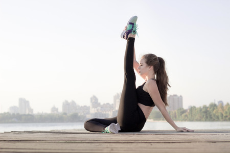 Young pretty slim fitness sporty woman stretches her legs during training workout exercises outdoor at river coast in the morningの写真素材