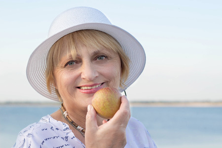 Happy retired elderly lady woman near lake shows white teeth biting appleの写真素材