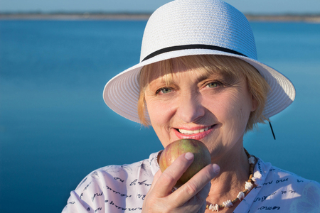 Happy retired elderly lady woman near lake shows white teeth biting appleの写真素材