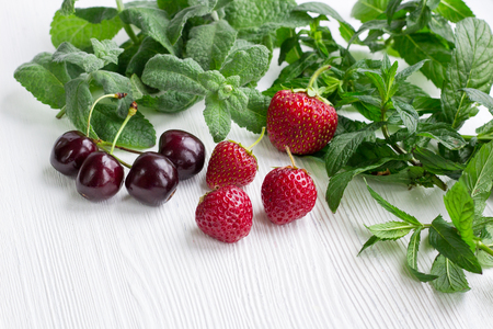 Vinous cherries, red strawberries with green herbal mix of fresh mint and melissa herbs on white wooden backgroundの写真素材