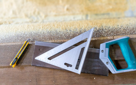 Carpentry equipment, including a hacksaw, square, and cutter, is seen in top view on a wooden table.