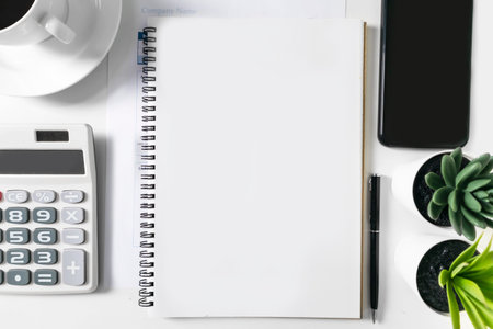 Blank white notebook with empty space in the middle. On the white table are a calculator and a mobile phone and a coffee cup, for mockup. Empty space for text, graphics work.の写真素材