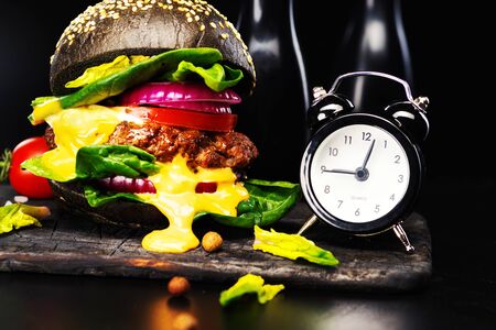Black beef burger with flowing melted cheese  and clock on a burnt blackboard  on a black background. Close-up.の写真素材