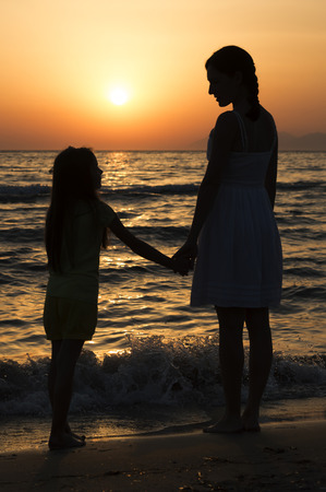 mother and daughter standing on sandy beach with waves breaking around their legs and looking at each other. Only dark silhouettes are visible. Bright red sunset can be seen in the background.の写真素材