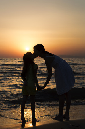 mother and daughter standing on sandy beach with waves breaking around their legs and kissing. Only dark silhouettes are visible. Bright red sunset can be seen in the background.の写真素材