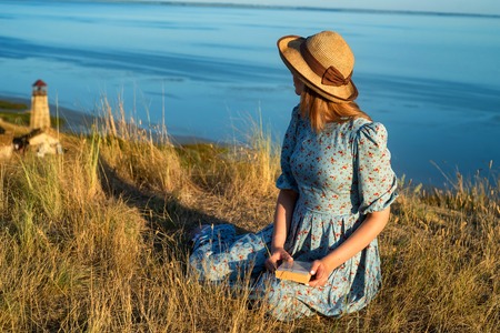 Romatic girl in dress and hat sits near seaの写真素材