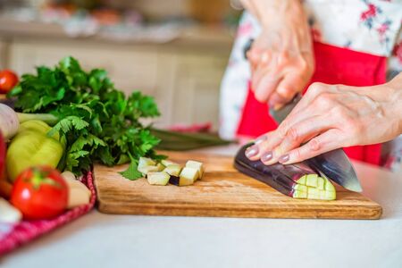 Close up female hand with knife cuts eggplant on wooden board in kitchen
