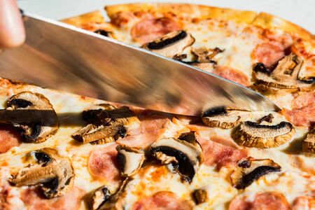 Close up knife slicing pizza with salami, mushrooms and cheese close upの写真素材
