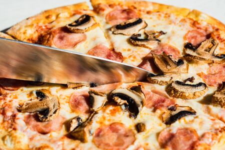 Close up knife slicing pizza with salami, mushrooms and cheese close upの写真素材