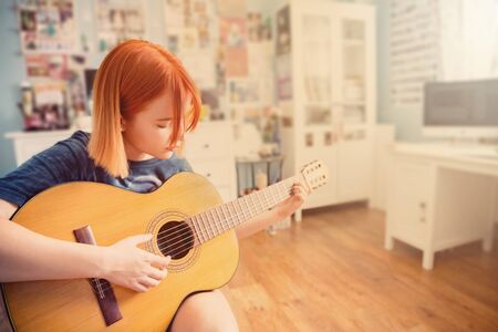 Pretty red-haired teenage girl plays guitar in her roomの写真素材