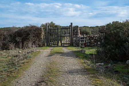 A rustic wooden gate with dry stone walls, located in a rural countryside with a dirt path and natural vegetation. The setting conveys tranquility and authenticityの写真素材