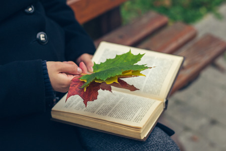 Girl in a black coat holding maple leaves and a book in the park in autumnの写真素材