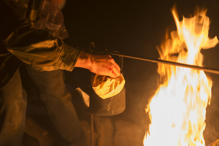 Hand of man taking the kettle, wich heated on a bright fire at night outdoor