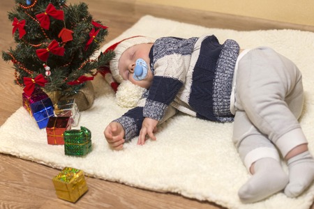 Little baby with nipple in Santa's hat sleeping under the Christmas tree with gifts